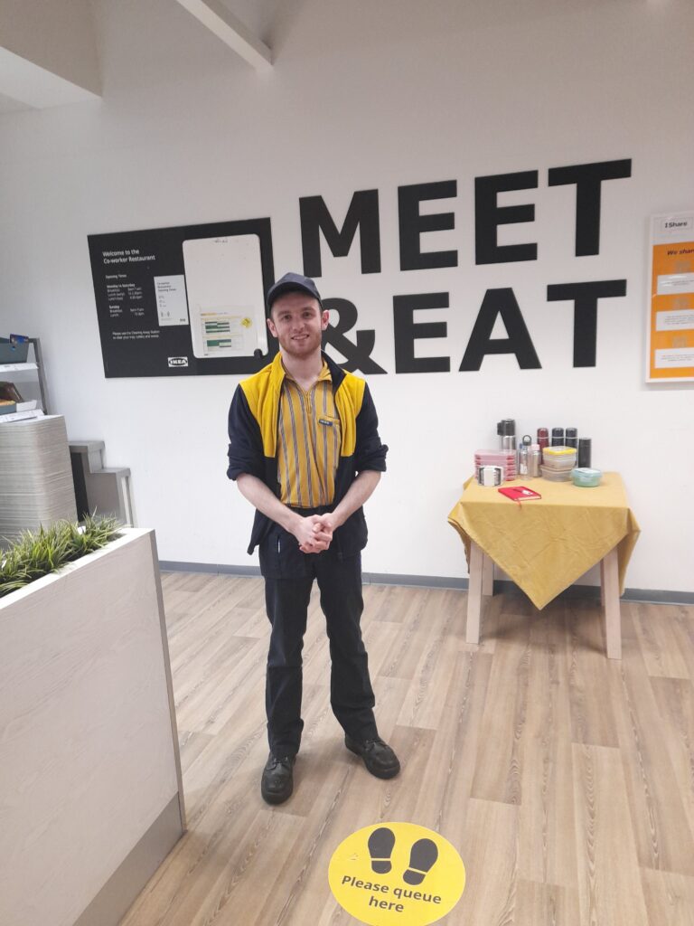Image of Daniel at work at Ikea stood in the canteen with his Yellow striped Ikea uniform on smiling at the camera