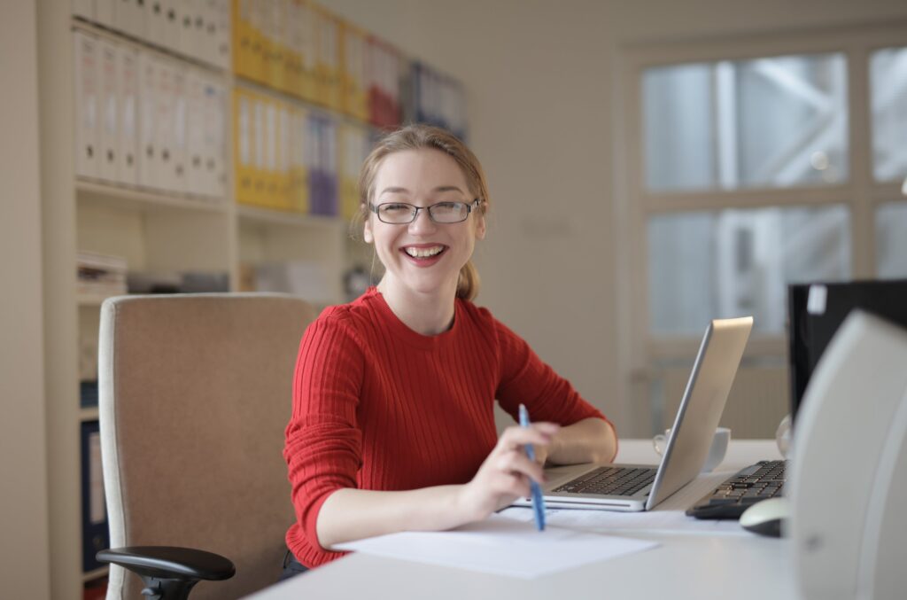 Image of lady sat at a desk with a laptop, holding a pen smiling at the camera