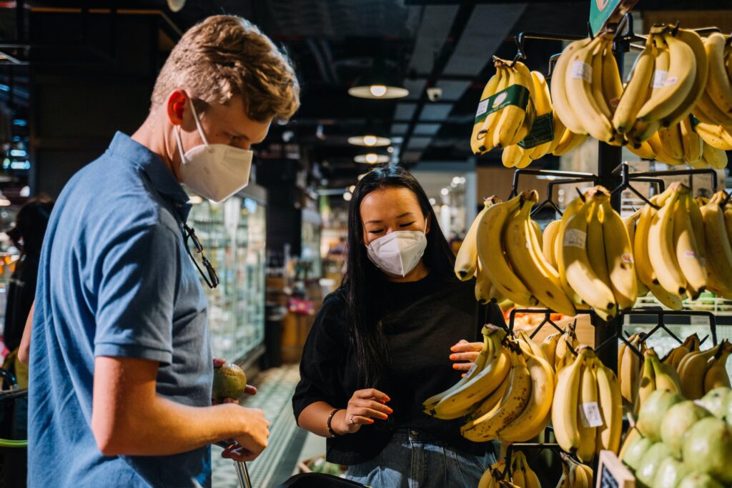 Image of a young man and a lady stood in a supermarket looking at bananas with masks on
